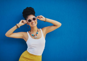 A person wearing colorful jewelry and round sunglasses beams at the camera while holding their sunglasses on their face. They stand against a deep blue backdrop.