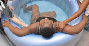 A pregnant person lies in a birthing pool, supported by two midwives.